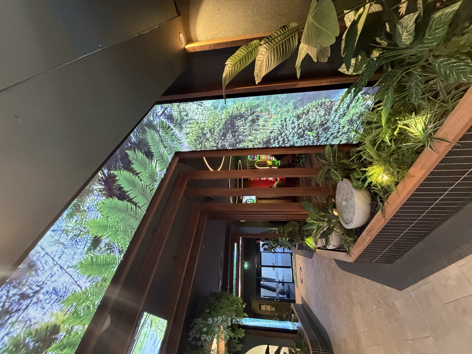 A wall of LED behind timber posts, ferns and tree trunks on it, with real plants in front